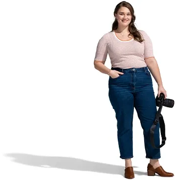 A woman in a tan pullover and blue jeans poses with her camera